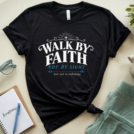 A black graphic t-shirt laid flat on a light beige surface, styled with a notebook, glasses, a plant, a mug, and folded mint-green shorts. The t-shirt features the text 'WALK BY FAITH NOT BY SIGHT' in white and light blue lettering.