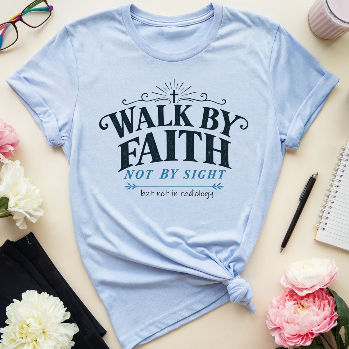 A light blue graphic T-shirt laid flat on a beige surface, surrounded by flowers, glasses, a drink, notebook, pen, and folded black fabric. The shirt has a printed slogan reading “WALK BY FAITH NOT BY SIGHT” with smaller text below.