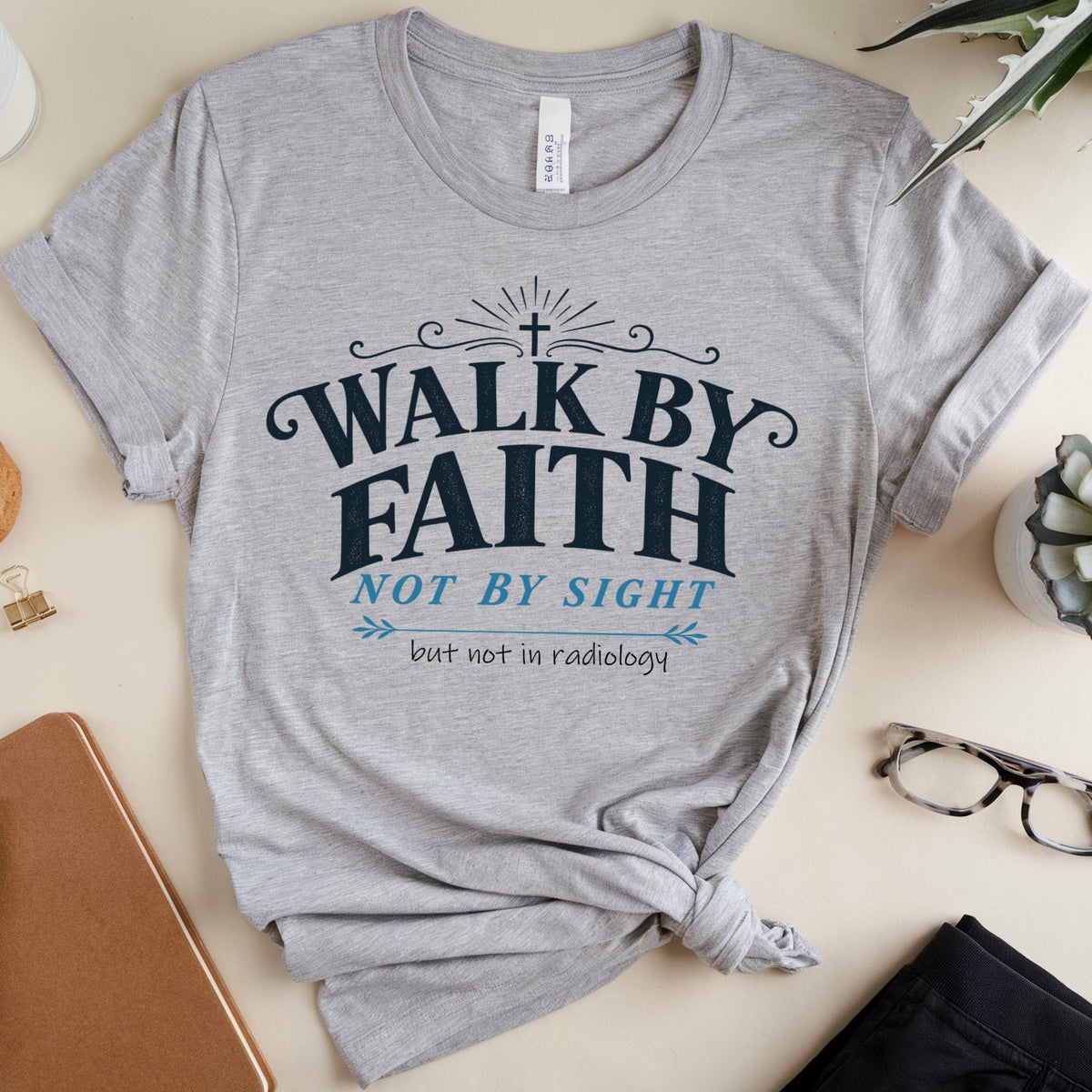 A heather gray graphic T-shirt laid flat on a beige surface, styled with a notebook, glasses, plant, cookie, milk glass, and folded pants. The shirt has a large front print reading “WALK BY FAITH NOT BY SIGHT but not in radiology” in dark blue and light blue text.