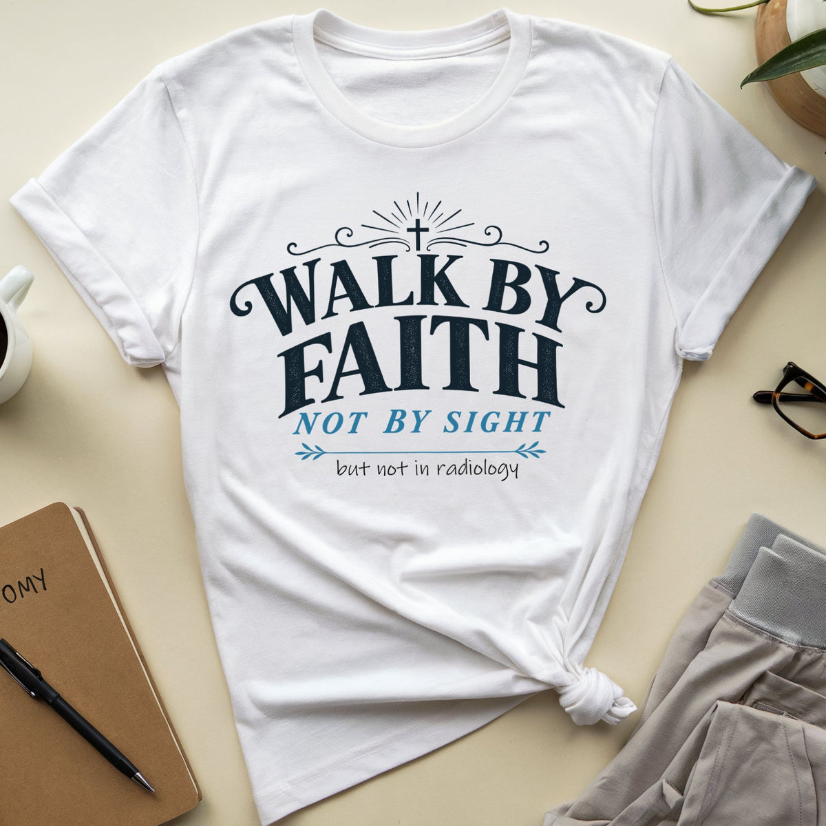 A white graphic T-shirt laid flat on a beige surface, styled with a coffee mug, notebook, glasses, folded pants, and a plant. The shirt has dark navy text reading “WALK BY FAITH NOT BY SIGHT” with smaller blue and black text below.