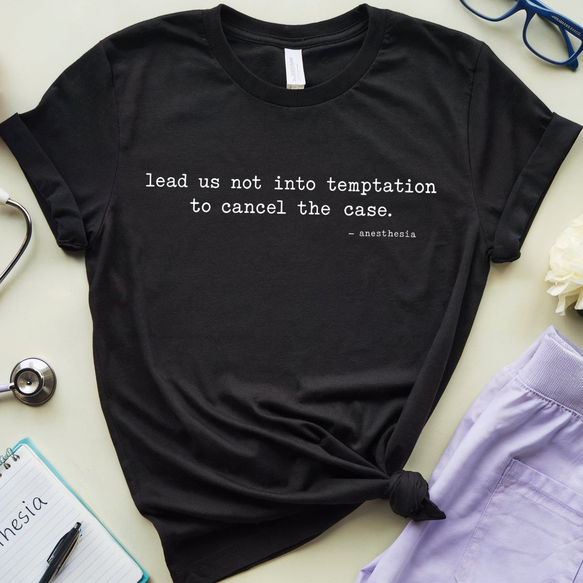 A black graphic t-shirt with a white quote printed on the front, styled in a flat lay with medical-themed props including a stethoscope, notebook, glasses, and lavender scrubs on a light beige background.