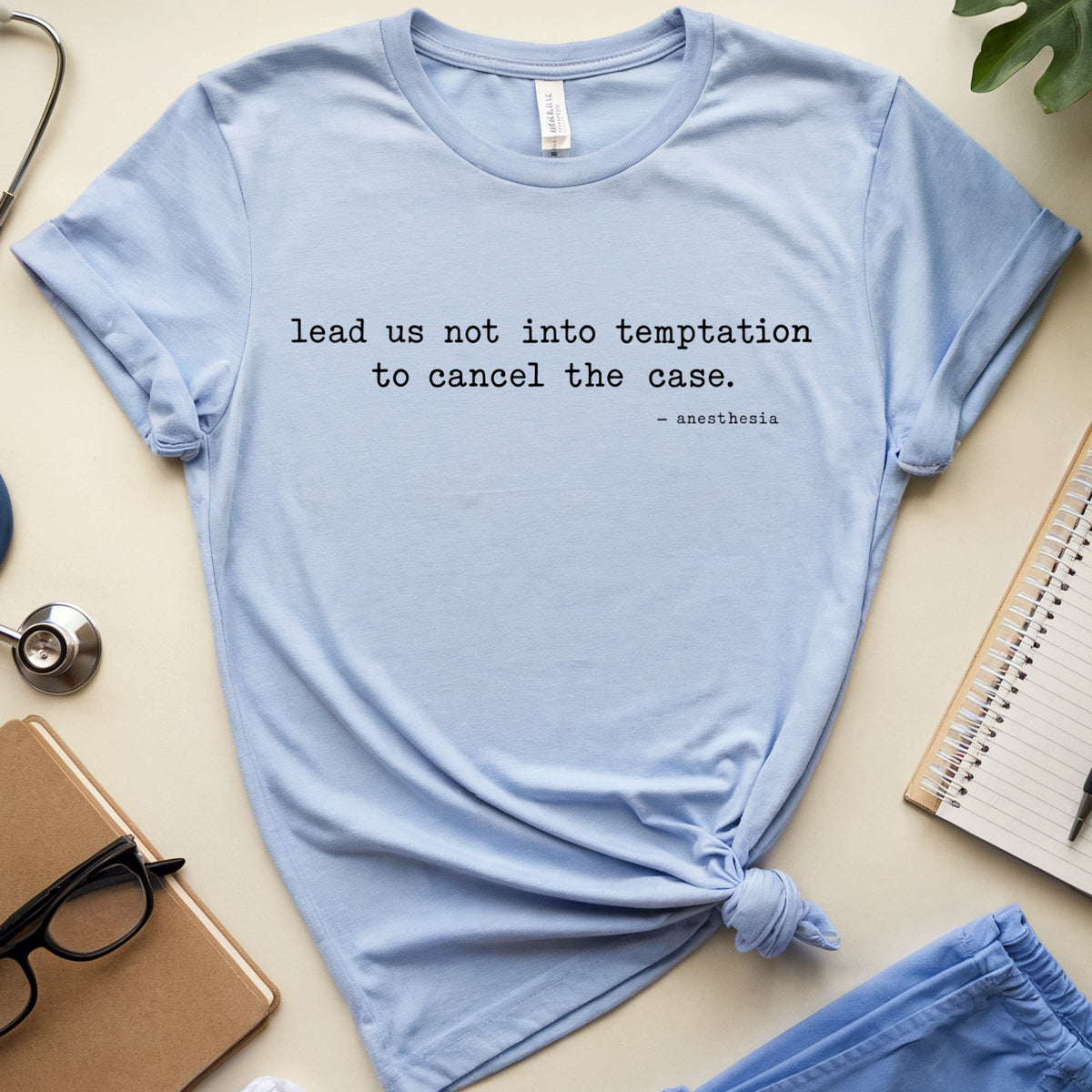 A light blue graphic T-shirt laid flat on a cream surface, styled with medical-themed props including a stethoscope, notebook, glasses, and folded scrubs. The shirt has black text reading 'lead us not into temptation to cancel the case.