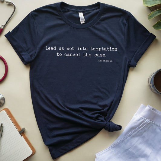 A navy blue graphic t-shirt with the quote 'lead us not into temptation to cancel the case.' in white text, styled in a flat lay with a stethoscope, notebook, plant, and coffee cup on a beige background.