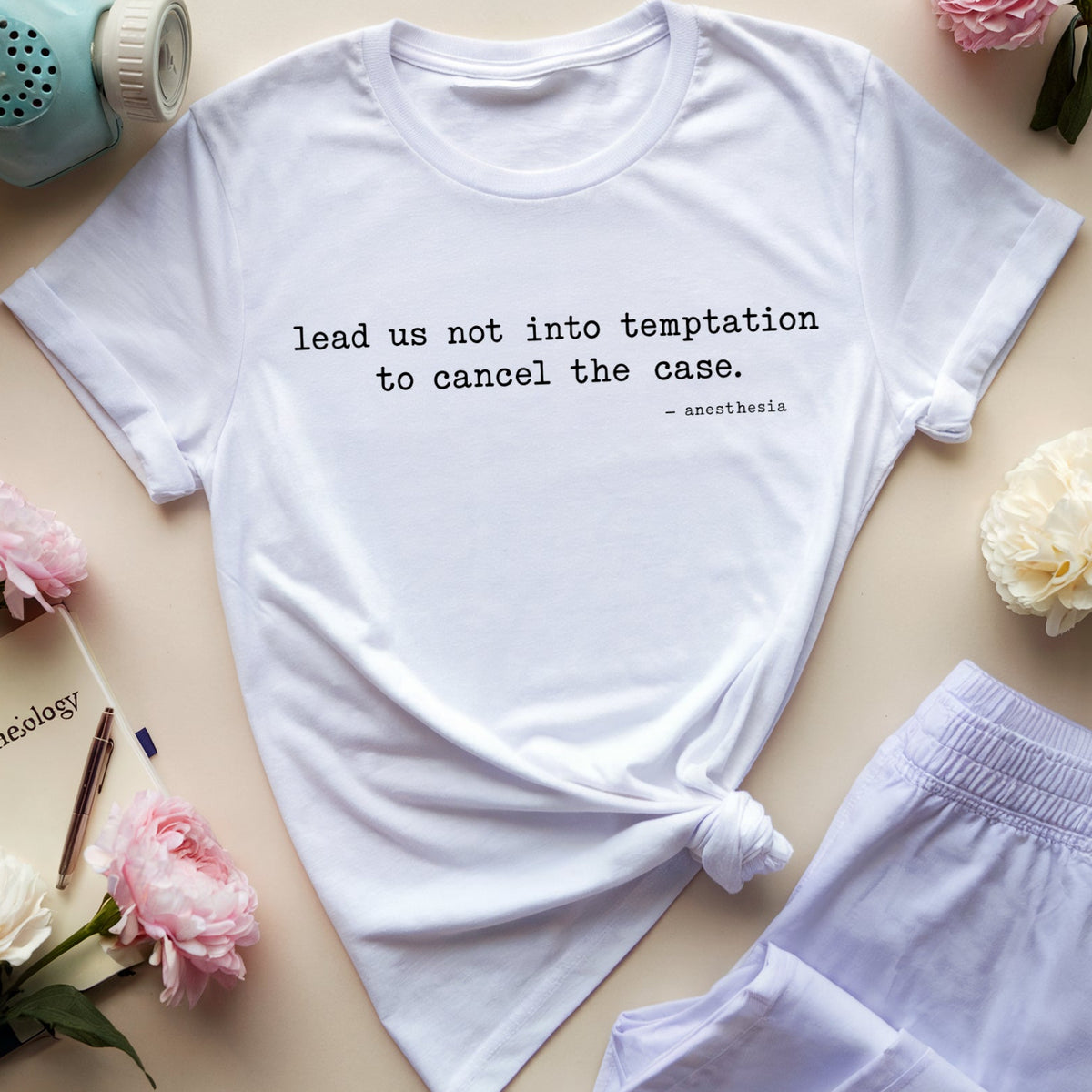 A white graphic T-shirt laid flat on a beige surface, styled with a light blue gas mask, purple eyeglasses, pink and white flowers, a book, and lavender shorts. The shirt has black text reading 'lead us not into temptation to cancel the case.
