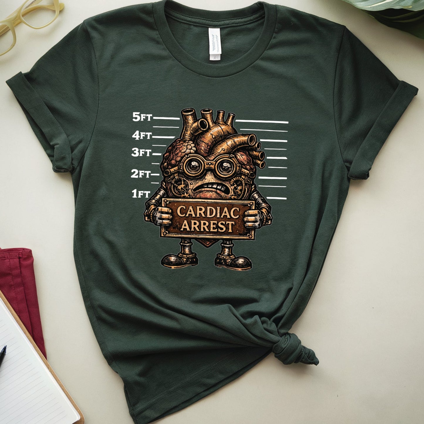 Green t-shirt with a graphic design of a heart with goggles and 'Cardiac Arrest' text, placed on a light surface with a plant and glasses in the background.