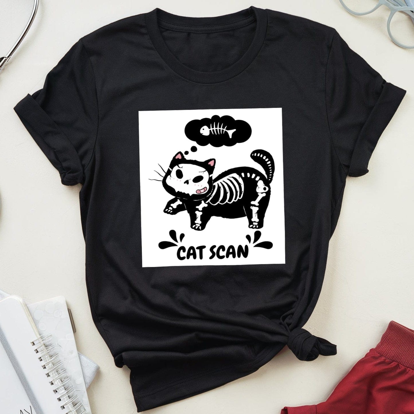Black t-shirt with a cat skeleton graphic and 'CAT SCAN' text on a light surface with medical-themed items.