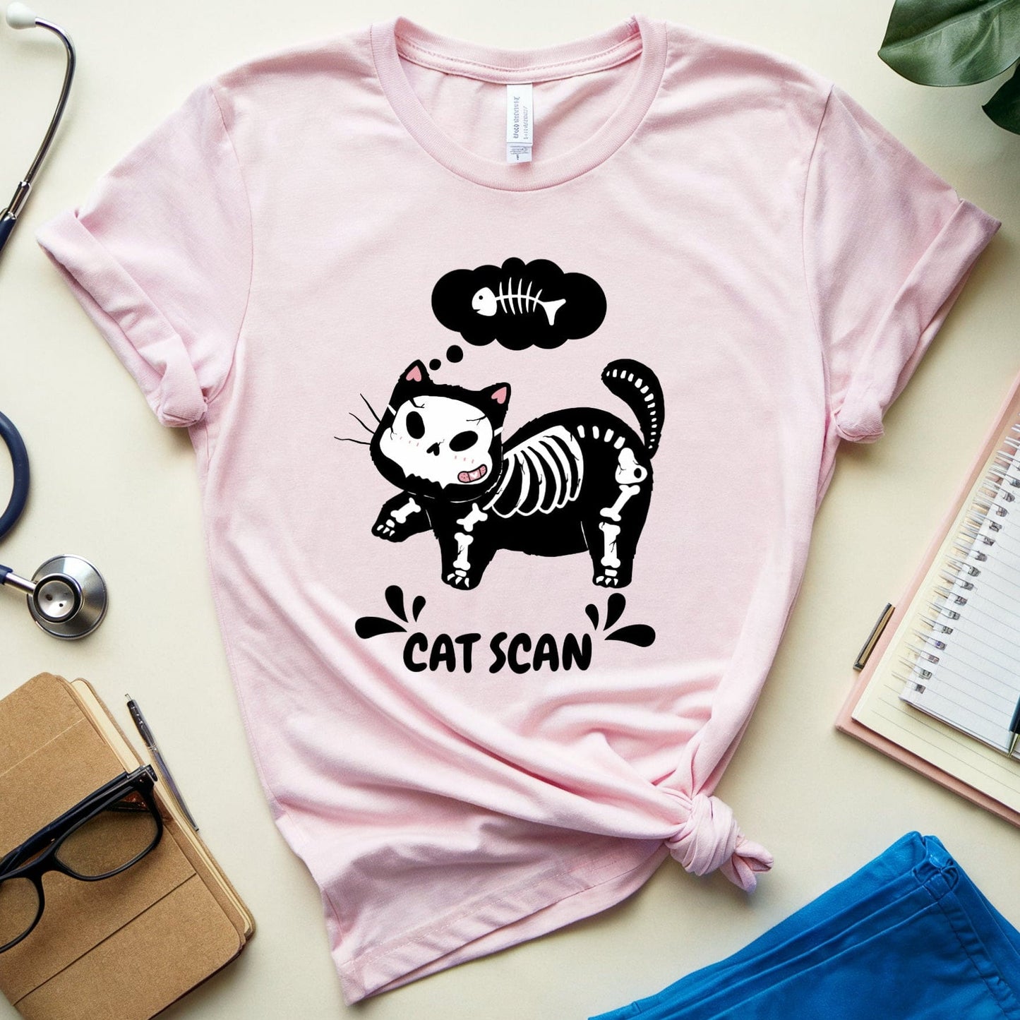 Pink t-shirt with a cat skeleton graphic and 'Cat Scan' text, surrounded by medical-themed items on a light surface.