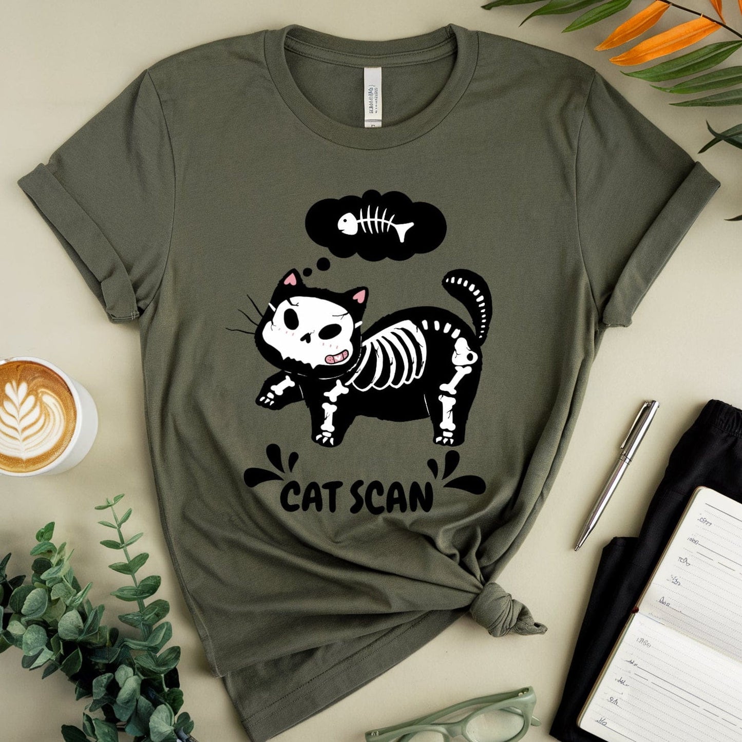 Green t-shirt with skeleton cat design, cup of coffee, eyeglasses, and notebook on a beige surface.