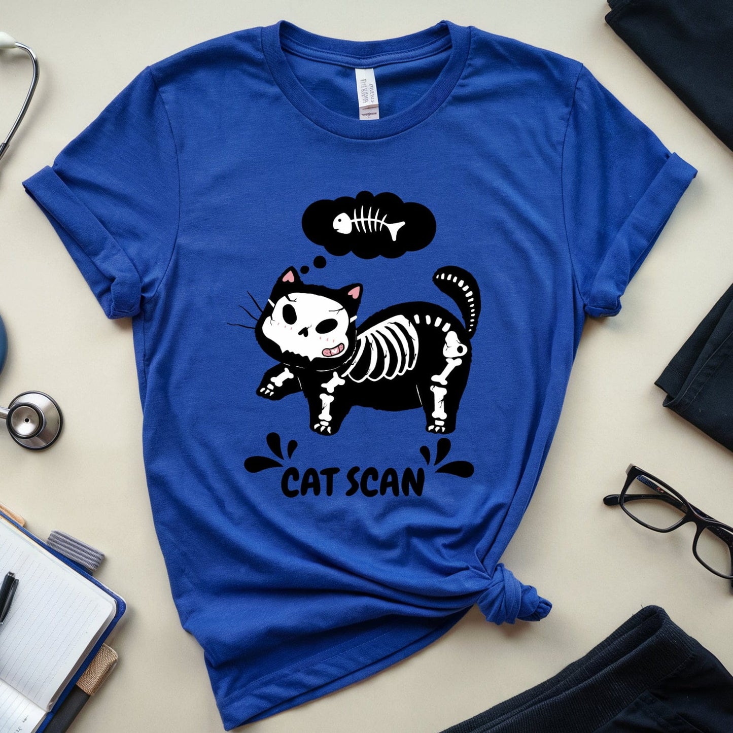 Blue t-shirt with a cat skeleton graphic and 'Cat Scan' text, surrounded by medical-themed items on a light surface.