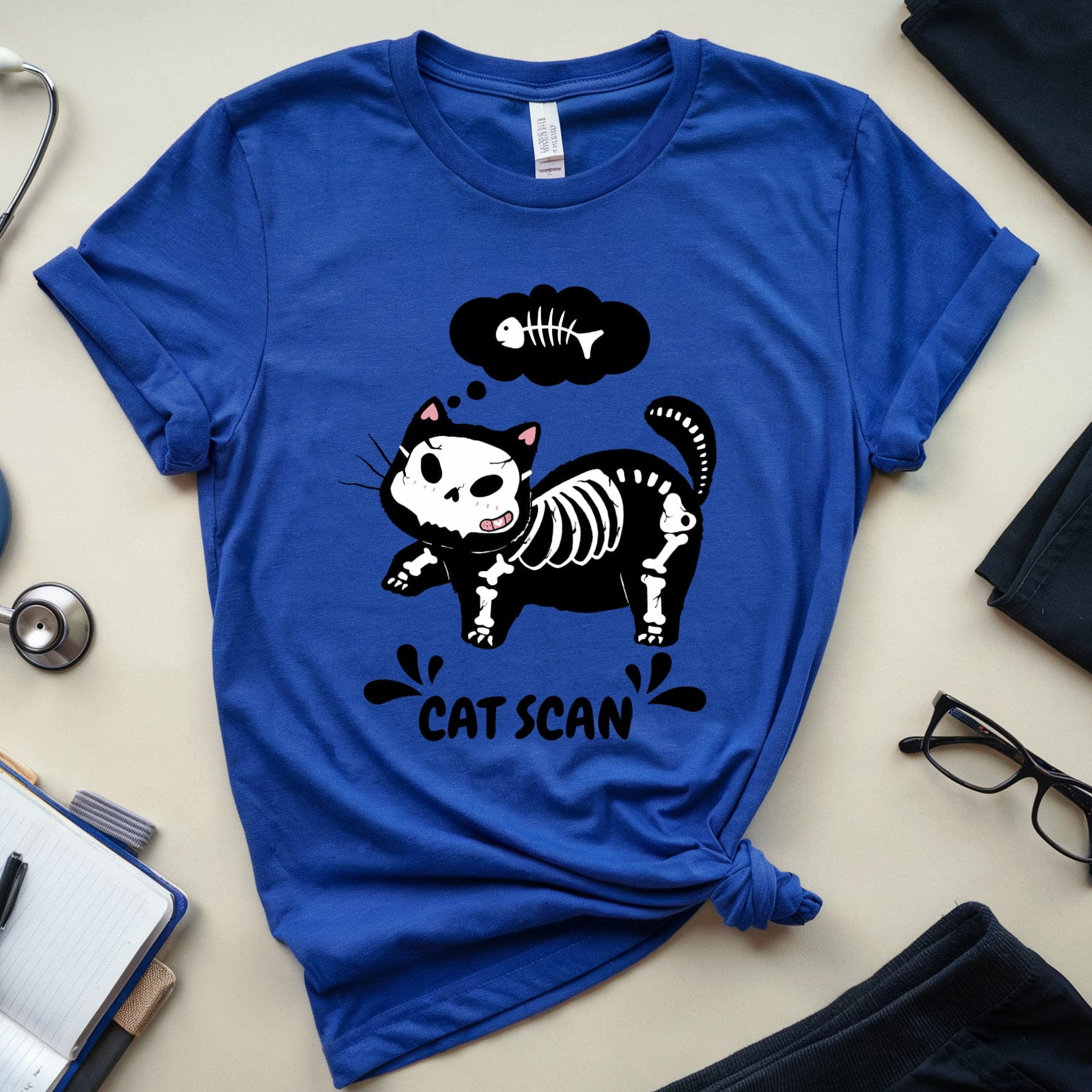 Blue t-shirt with a cat skeleton graphic and 'Cat Scan' text, surrounded by medical-themed items on a light surface.