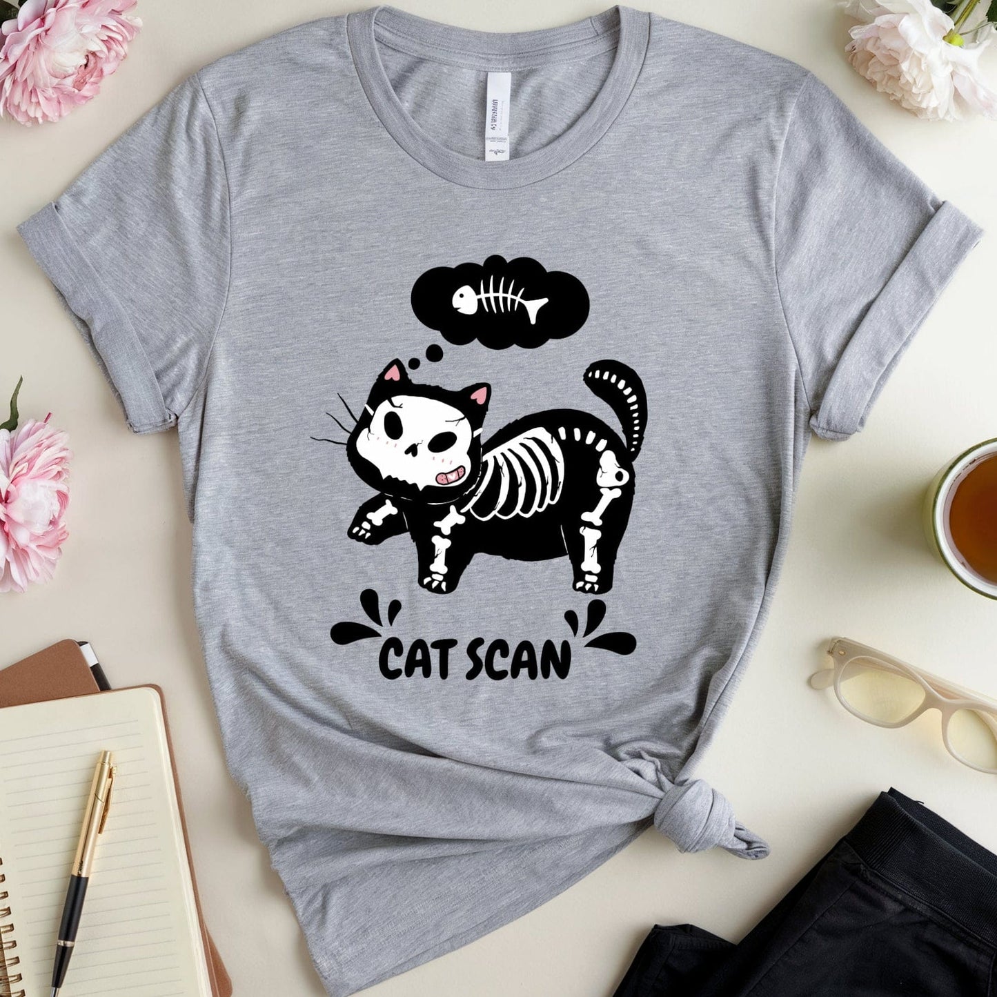 Gray t-shirt with a black cat skeleton graphic and 'Cat Scan' text on a light surface with flowers and a cup.