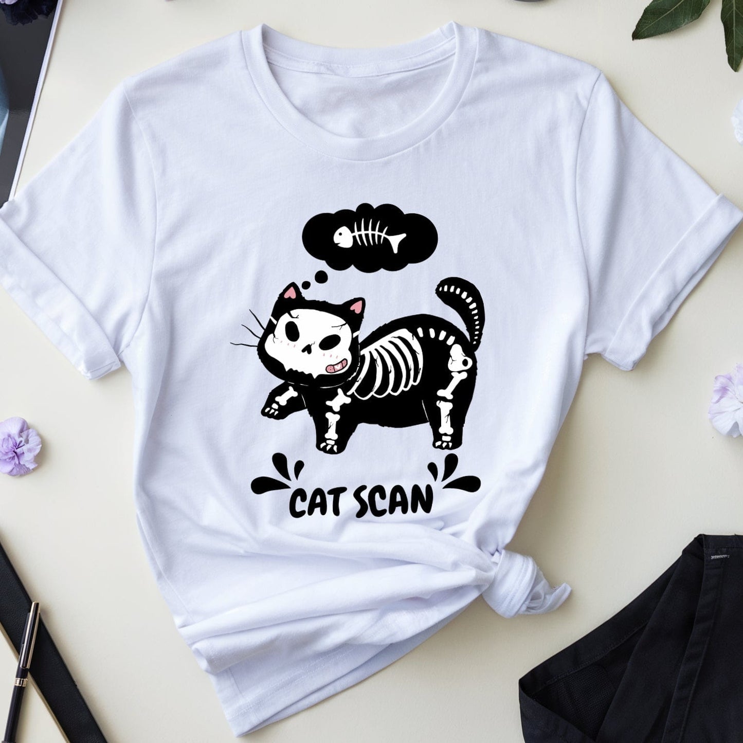 White t-shirt with a cat skeleton graphic on a light surface with medical-themed items.