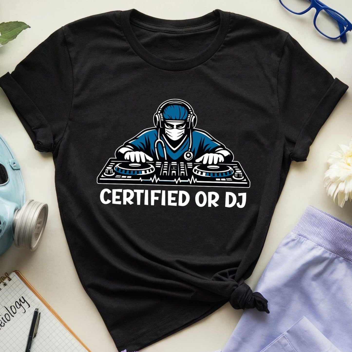Black t-shirt with a graphic design of a person on DJ equipment and 'CERTIFIED OR DJ' text, surrounded by flowers and accessories on a light surface.
