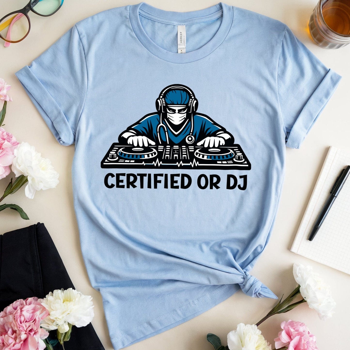 Light blue t-shirt with a graphic of a person on DJ equipment and 'CERTIFIED OR DJ' text, placed on a surface with flowers and glasses.