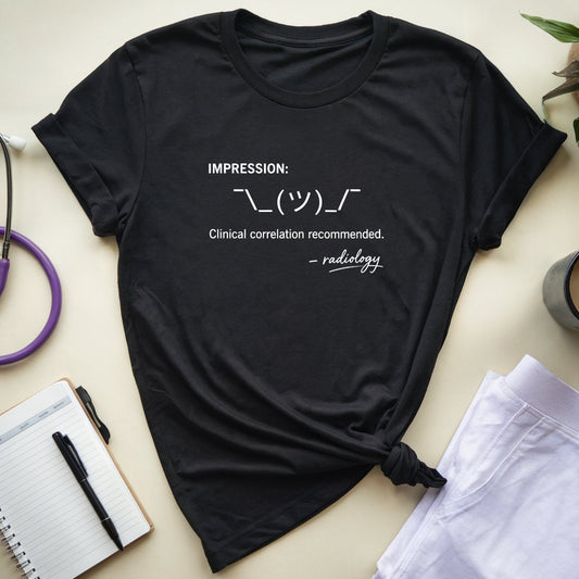 A black short-sleeve graphic T-shirt laid flat on a light beige surface, styled with a stethoscope, notebook, pen, folded white pants, a mug, and a plant. The shirt has white printed text reading 'IMPRESSION: ”··(м)··” and 'Clinical correlation recommended.