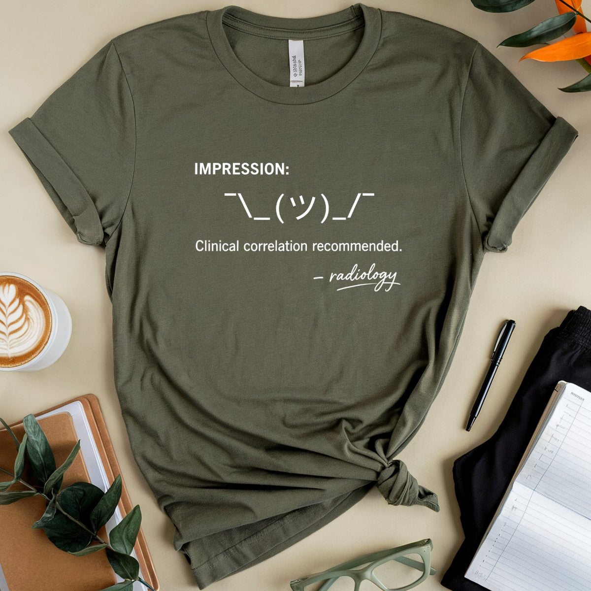 An olive green graphic t-shirt is styled in a flat lay on a beige background. The shirt features white text reading 'IMPRESSION: ” followed by a radiology symbol and 'Clinical correlation recommended.