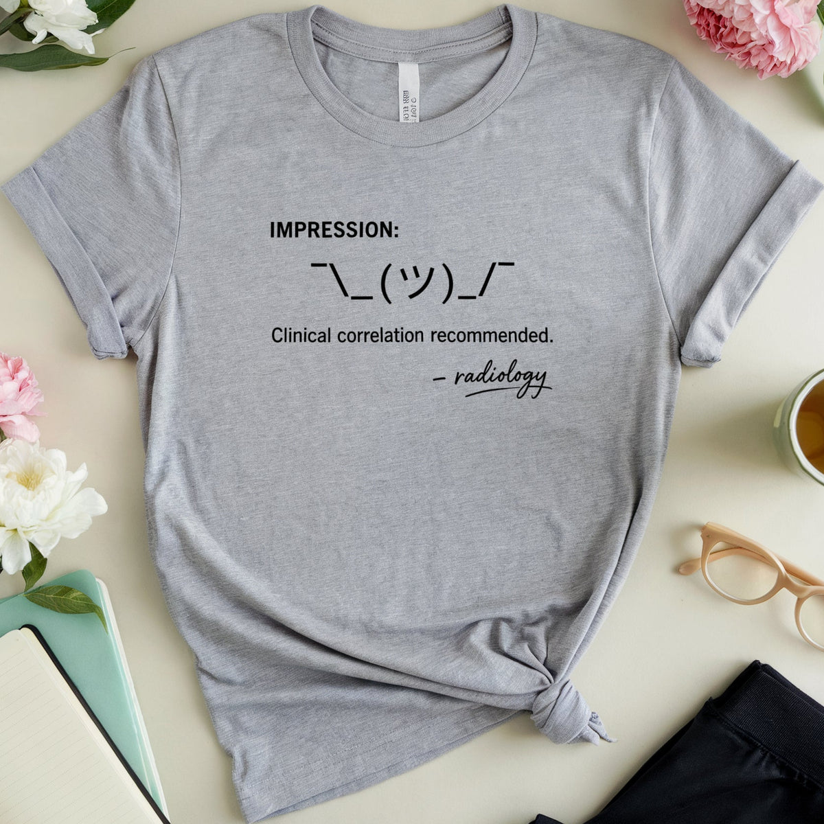 A heather gray graphic T-shirt laid flat on a light beige surface with flowers, a notebook, glasses, a cup of tea, and folded pants around it. The shirt has black printed text reading 'IMPRESSION: ❤️❤️ Clinical correlation recommended.