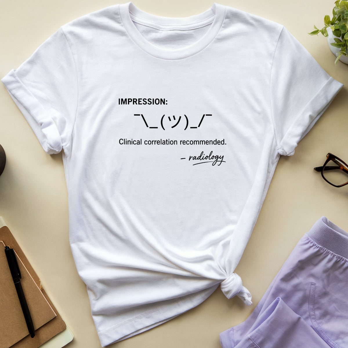 A white short-sleeve graphic T-shirt laid flat on a light beige surface, styled with a notebook, coffee cup, eyeglasses, and lavender shorts. The shirt has black printed text reading 'IMPRESSION: \_(\_)_/ Clinical correlation recommended.