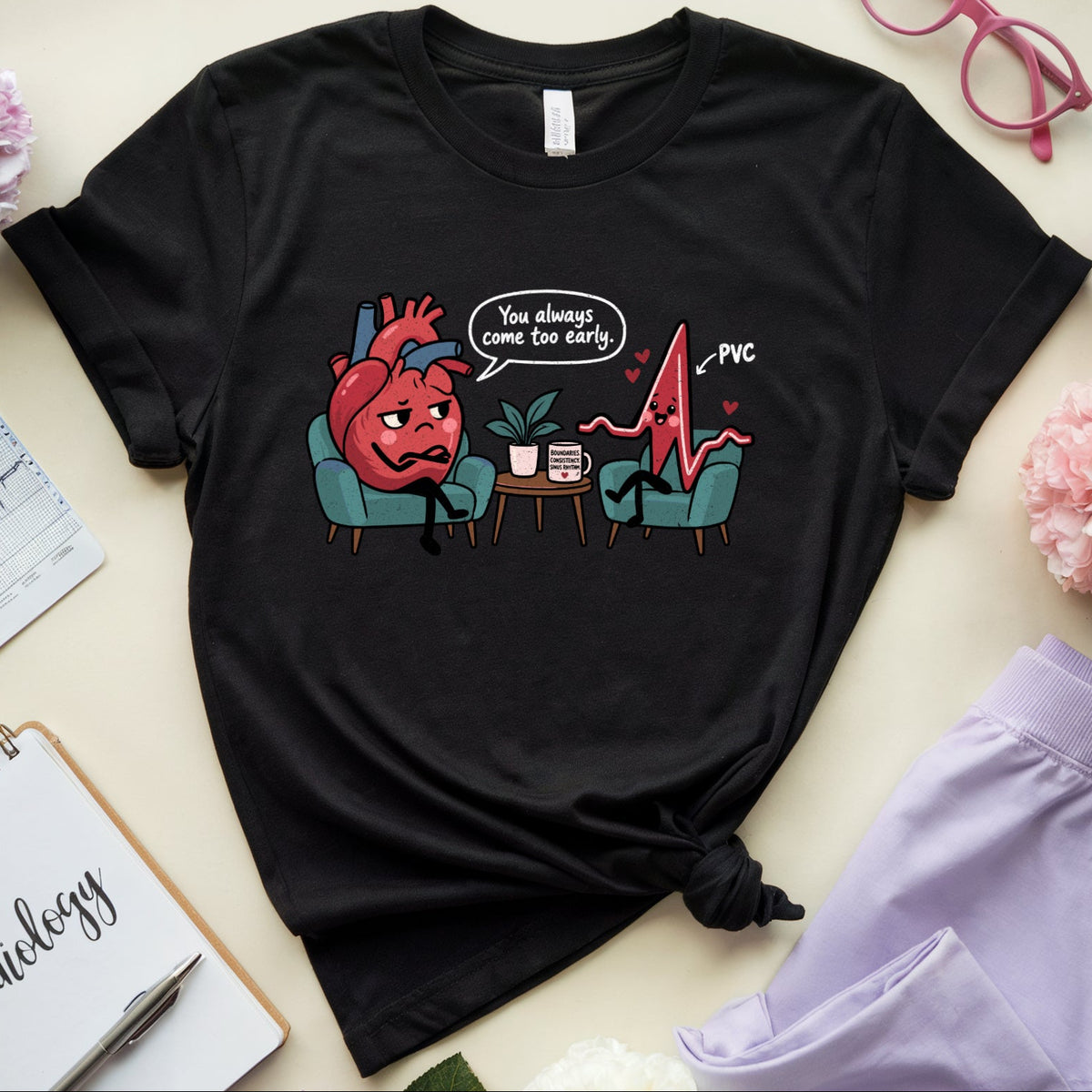 A black graphic t-shirt featuring a cartoon illustration of a red heart and a pink EKG line sitting in chairs, with the text 'You always see the heart. PVC'.
