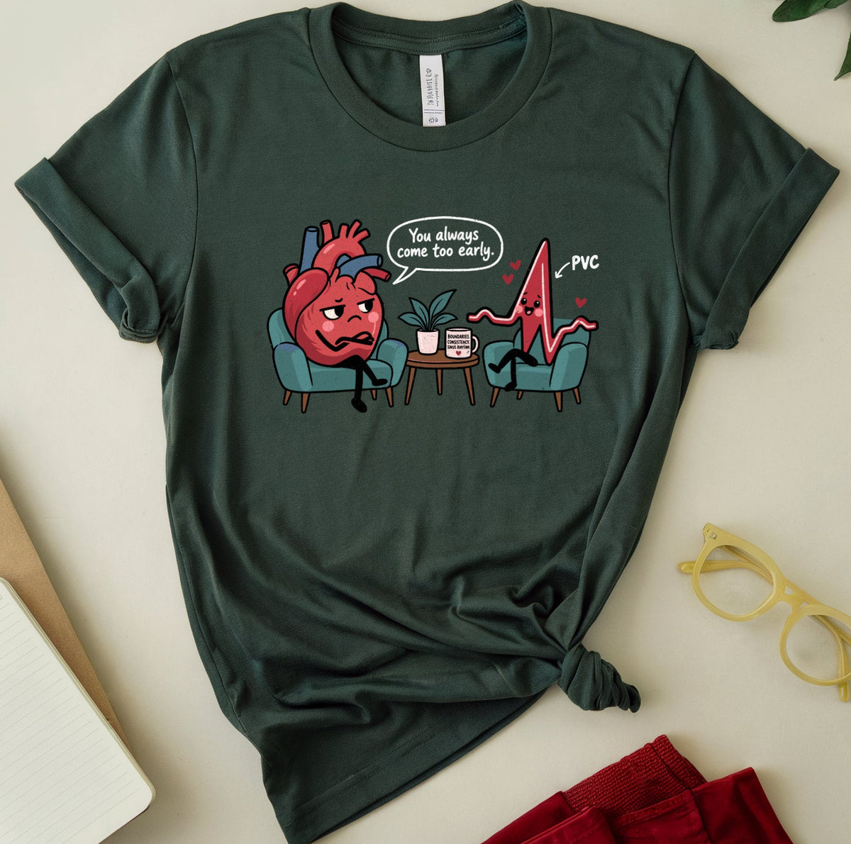 A dark green graphic t-shirt is displayed in a styled flat-lay arrangement on a light beige surface. The t-shirt features a cartoon graphic of a red heart sitting in a teal chair next to a red EKG line, with a speech bubble that reads 'I'm always calm for heart' and 'PVC'.