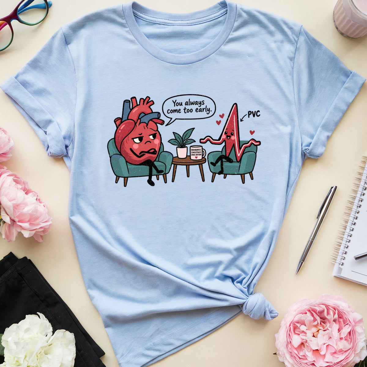 A light blue graphic t-shirt featuring a cartoon of a stressed-out red heart in an armchair next to a shocked pink heart with a speech bubble that reads 'You always come too late! -PVC'. The t-shirt is styled in a flat-lay arrangement with pink flowers, glasses, a notebook, and a drink on a beige background.