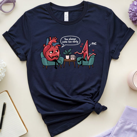 A navy blue graphic t-shirt is styled in a flat lay on a light beige background. The t-shirt features a cartoon graphic of a red heart sitting in a teal chair, looking distressed, with a speech bubble that reads 'You almost got me live right?' and a heartbeat line ending in 'PMS'.