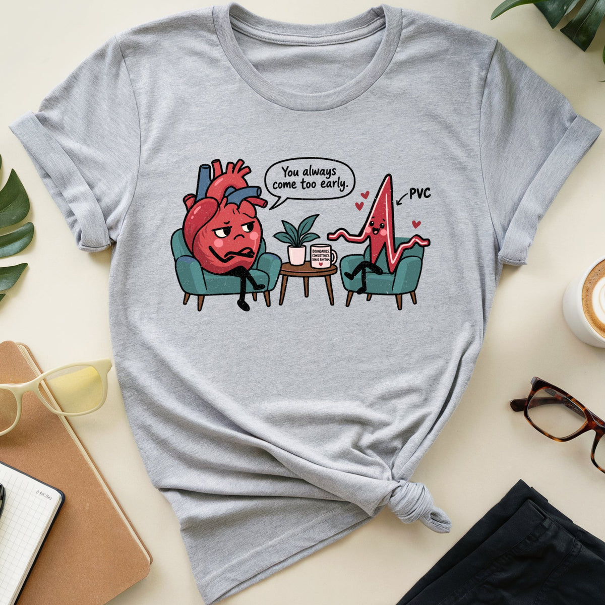 A light heather gray graphic t-shirt is displayed in a flat-lay arrangement on a cream-colored surface. The t-shirt features a cartoon graphic of a distressed red heart sitting in a teal chair next to a jagged red line labeled 'PVC' with a speech bubble that reads 'You always come too late.