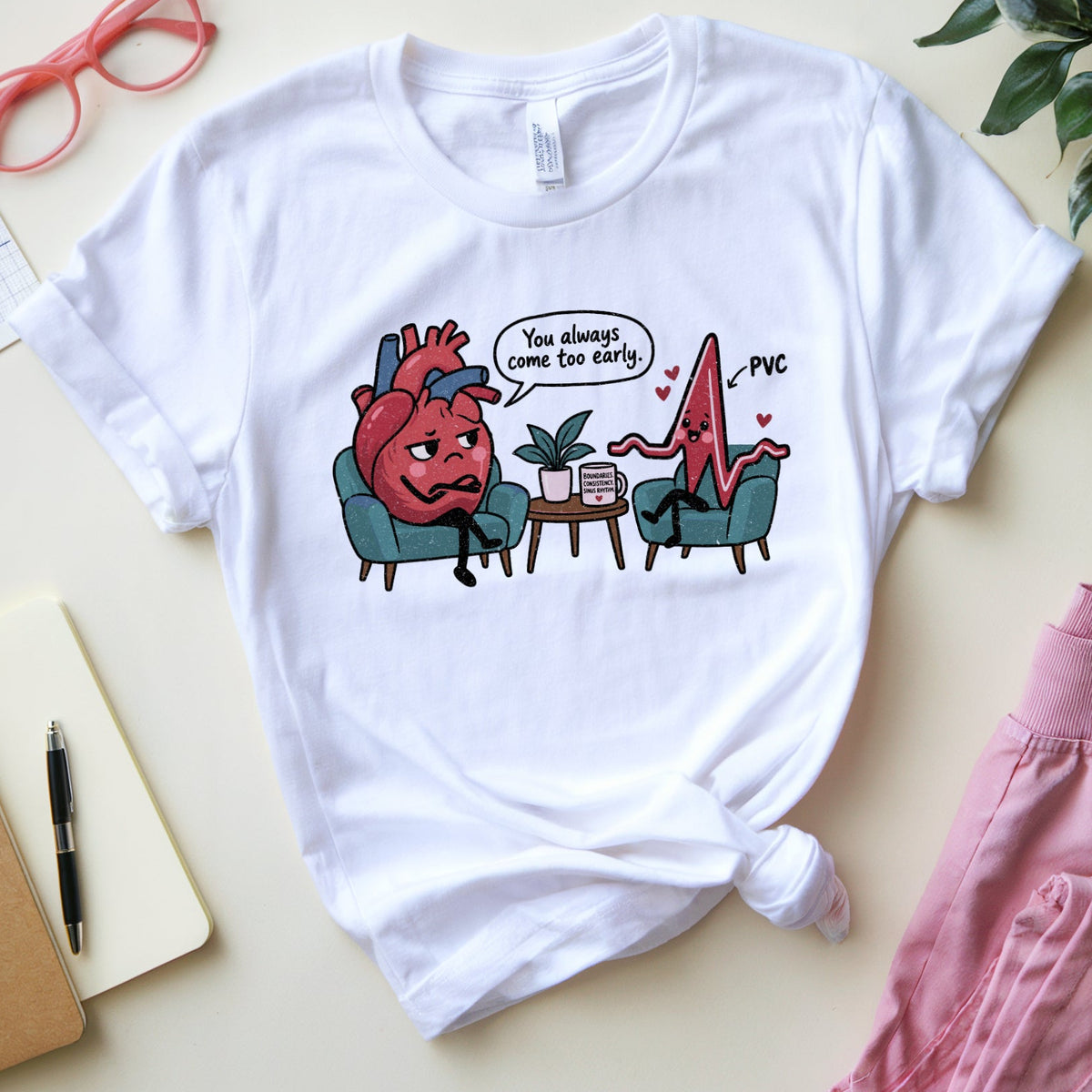 A white graphic t-shirt featuring a cartoon heart and blood pressure line sitting in chairs with a speech bubble reading 'You always come too early', styled in a flat lay with medical-themed props like a notebook, glasses, and a plant.