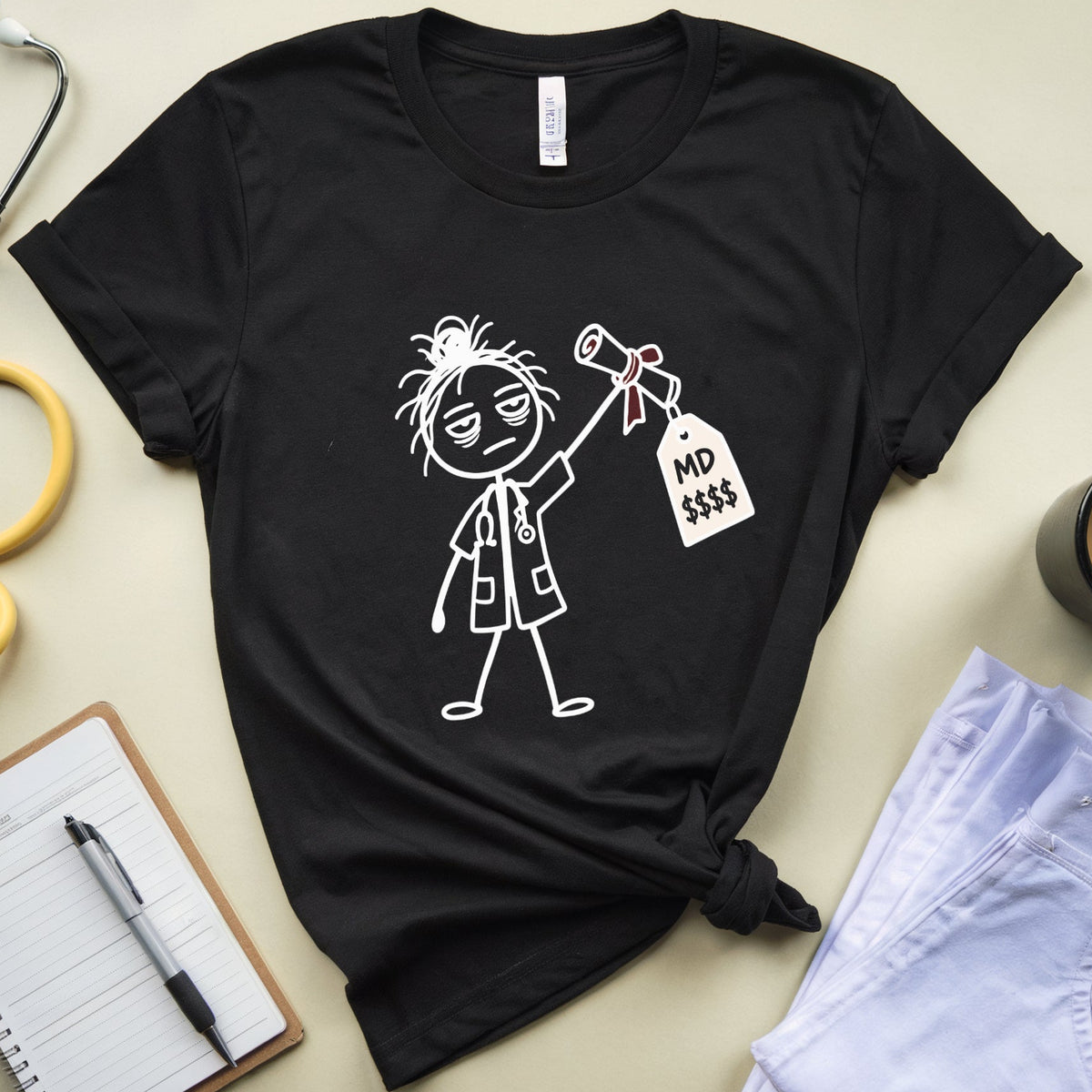 A black graphic t-shirt with a white line art illustration of a doctor holding a '$D' tag, styled in a flat-lay with a stethoscope, notebook, pen, coffee mug, and light blue jeans on a beige background.