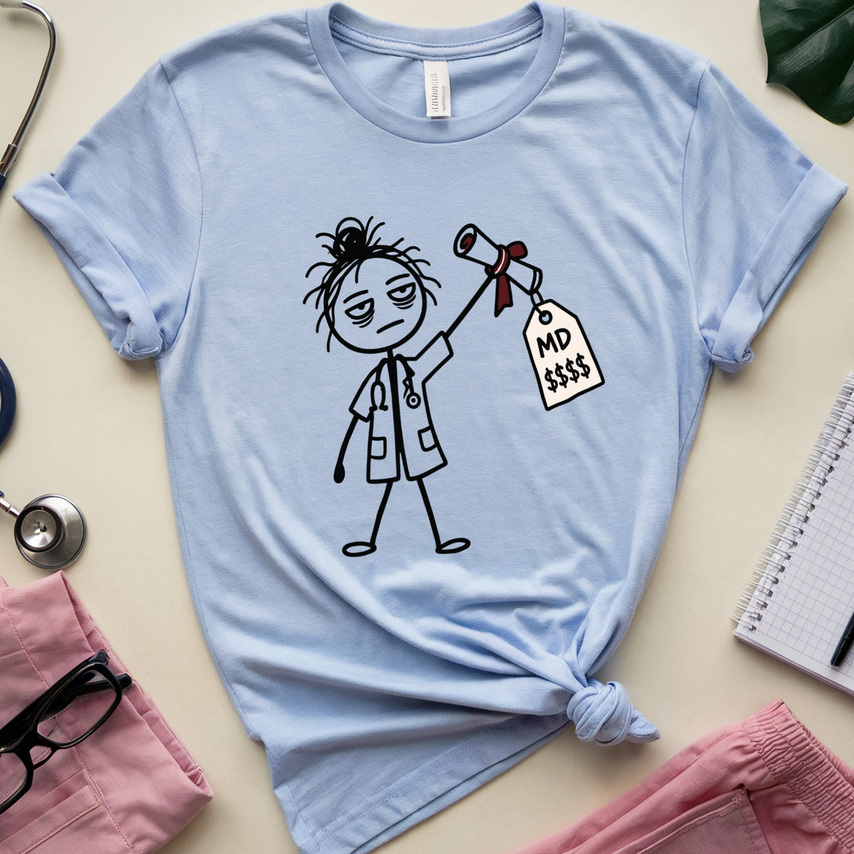 A light blue graphic t-shirt featuring a stick figure doctor holding a gavel with a price tag that reads 'NO £$$'. The t-shirt is styled in a flat lay with pink scrubs, a stethoscope, glasses, a notebook, and a green leaf on a beige background.