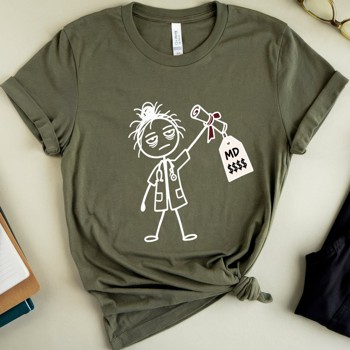 An olive green graphic t-shirt with a white line art drawing of a doctor holding a price tag that reads 'MD $$$'. The t-shirt is styled in a flat-lay arrangement on a light beige background, surrounded by props including eyeglasses, a notebook, a pen, and a pair of pants.