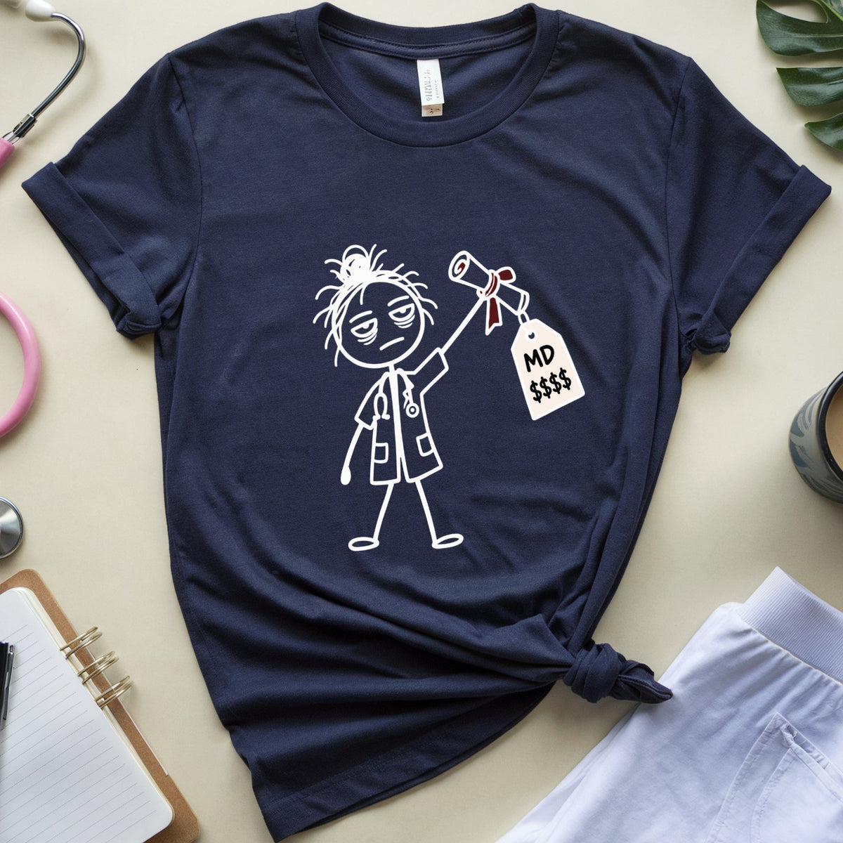A navy blue graphic t-shirt featuring a white line art illustration of a doctor holding a '$D $$$' price tag, styled in a flat lay with medical-themed props including a pink stethoscope, a notebook, and white scrubs on a beige background.