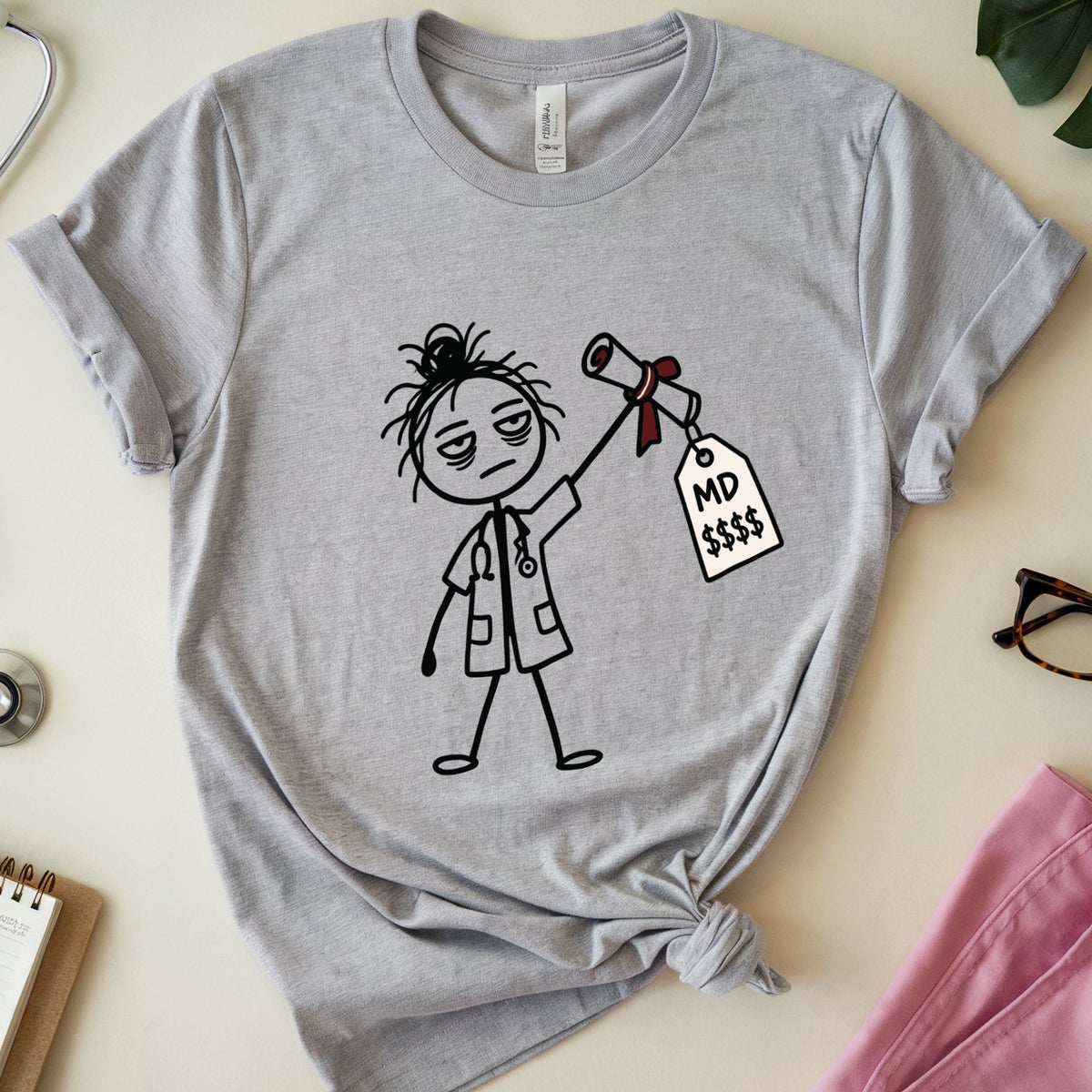 A heather gray graphic t-shirt featuring a cartoon doctor holding an MD tag, styled in a flat lay with medical props including a stethoscope, notebook, glasses, and pink scrubs on a beige background.