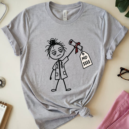 A heather gray graphic t-shirt featuring a cartoon doctor holding an MD tag, styled in a flat lay with medical props including a stethoscope, notebook, glasses, and pink scrubs on a beige background.