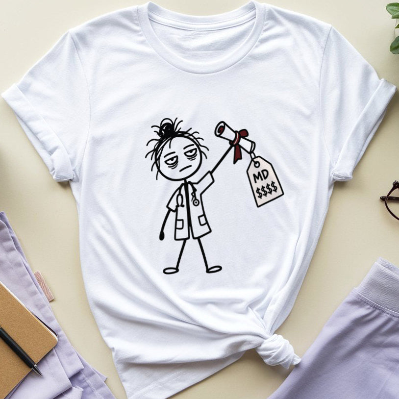 A white graphic t-shirt featuring a black line art illustration of a nurse holding a price tag that reads 'NO $$$'. The t-shirt is styled in a flat-lay arrangement on a light beige surface, surrounded by lavender scrubs pants, a notebook, glasses, a plant, and a coffee mug.