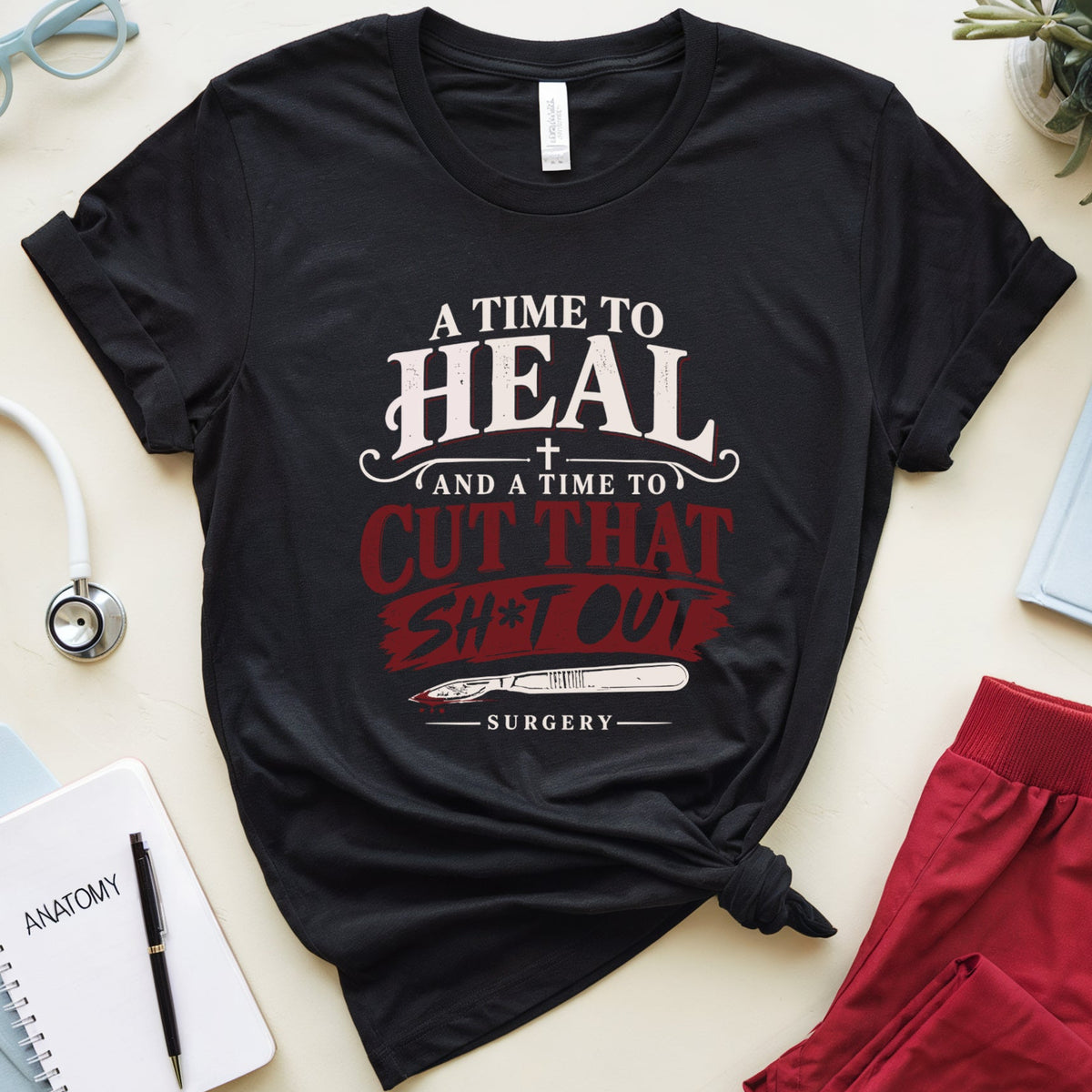 A black graphic t-shirt with a tie knot, featuring the text 'A TIME TO HEAL AND A TIME TO CUT THAT SH*T OUT SURGERY' in white and red. The shirt is styled in a flat lay with medical-themed props including red scrubs, a stethoscope, glasses, and notebooks on a light beige background.