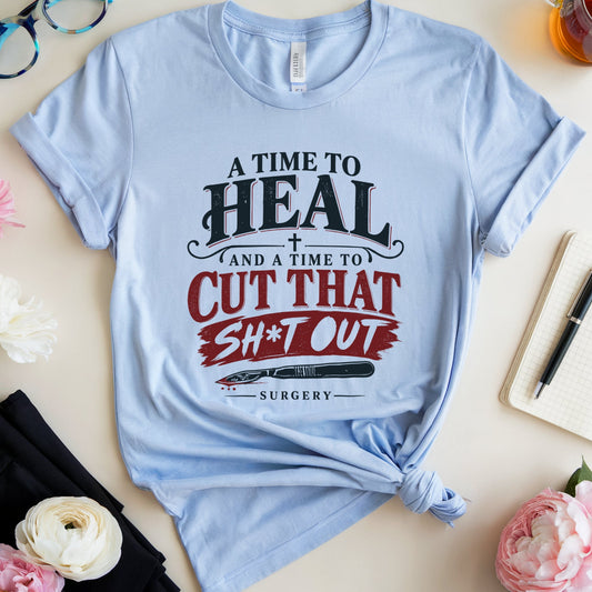 A light blue graphic t-shirt with a knotted hem, featuring the text 'A TIME TO HEAL (AND A TIME TO CUT THAT SH*T OUT) SURGERY' in black and red lettering. The t-shirt is styled in a flat-lay arrangement with pink and white flowers, black pants, blue-framed glasses, a notebook, a pen, and a glass of tea on a light beige background.
