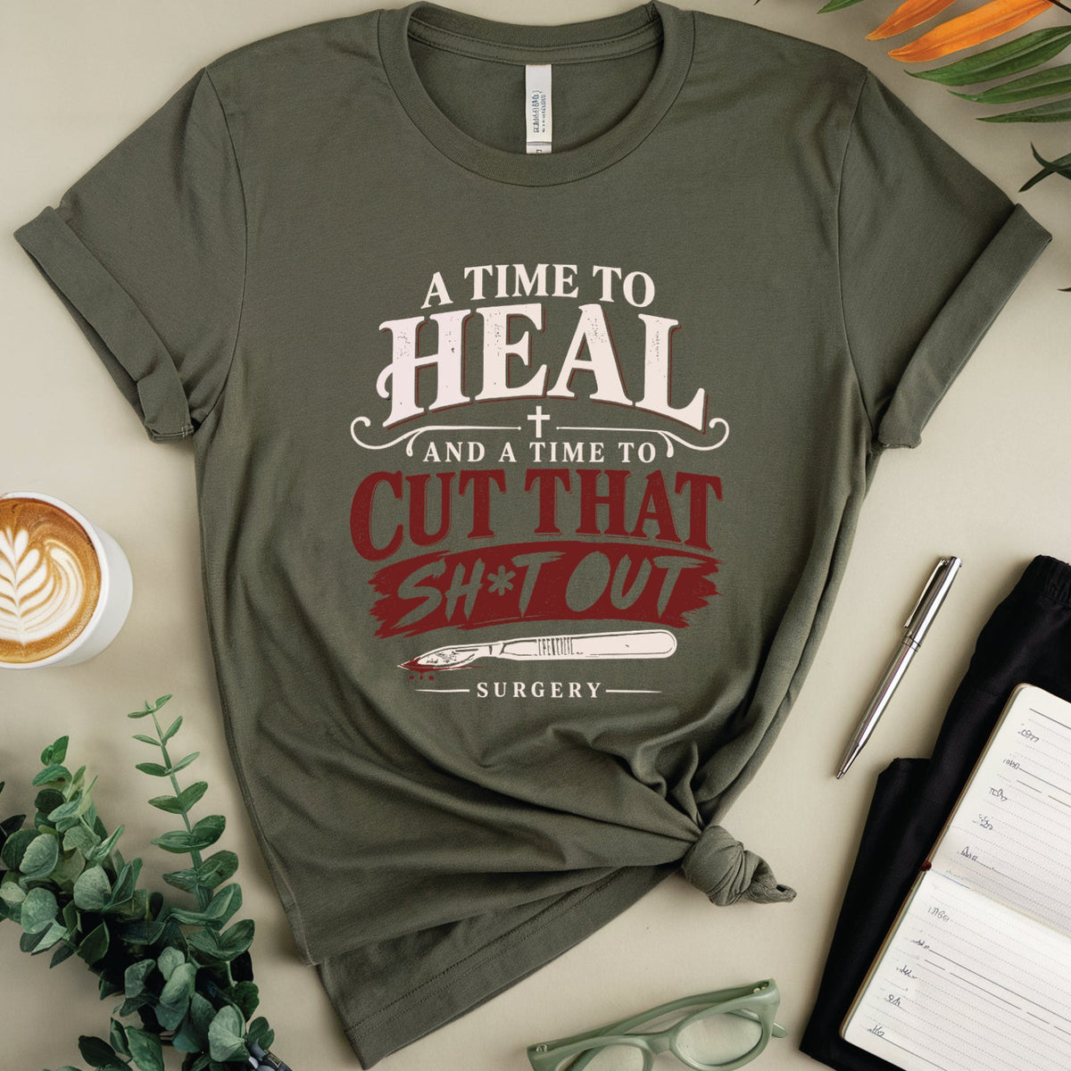An olive green graphic t-shirt with a humorous surgery-themed print, styled in a flat lay with a coffee cup, notebook, eyeglasses, and greenery on a beige background.