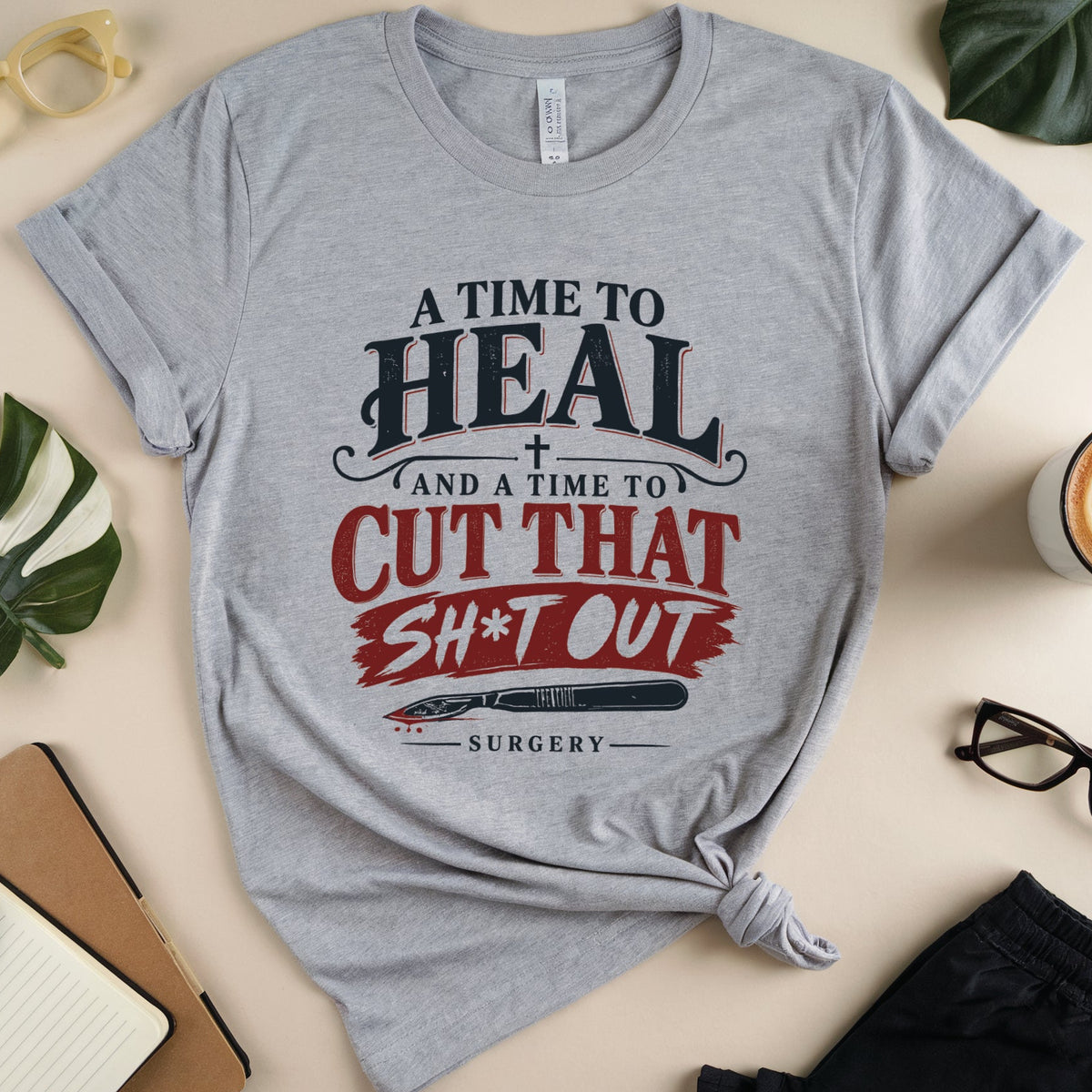 A heather gray graphic t-shirt with a 'Surgery' themed text print, styled in a flat lay with glasses, a coffee cup, a notebook, and plant leaves on a beige background.