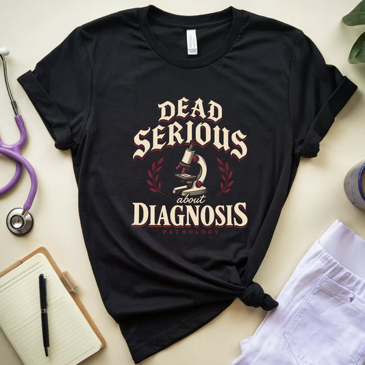 A black graphic T-shirt laid flat on a light beige surface, styled with a purple stethoscope, notebook, pen, plant leaves, a mug, and white pants. The shirt has a large cream and red text graphic reading 'Dead Serious About Diagnosis' with a microscope illustration and the word 'Pathology' below.
