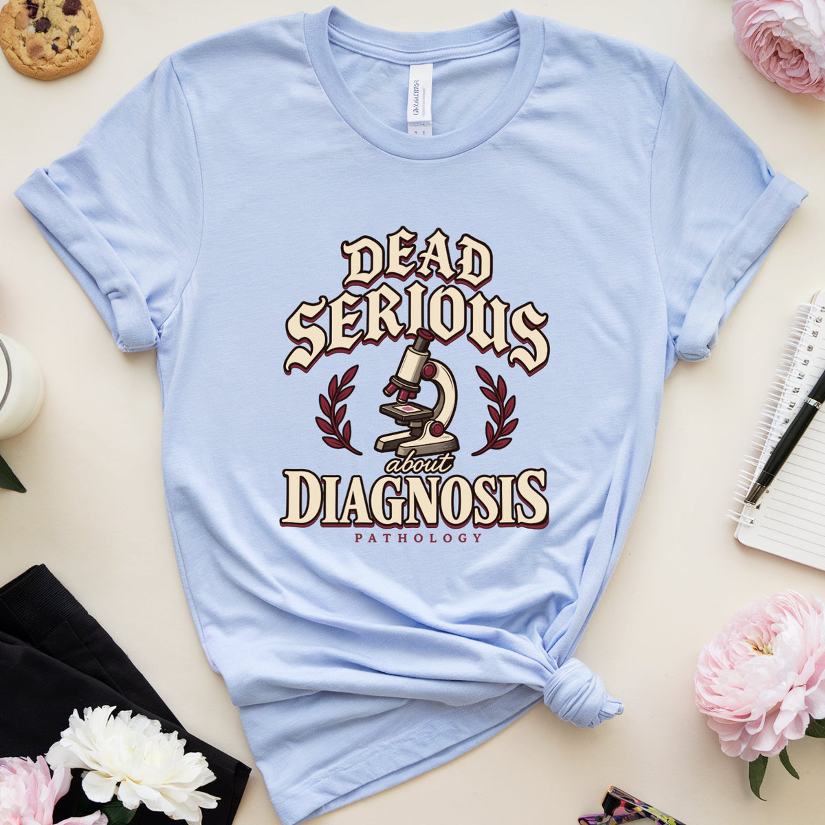 A light blue graphic t-shirt with a knotted hem, featuring the text 'DEAD SERIOUS ABOUT DIAGNOSIS' and a microscope graphic. The t-shirt is styled in a flat-lay arrangement with pink flowers, a cookie, a notebook, glasses, and a glass of milk on a beige background.