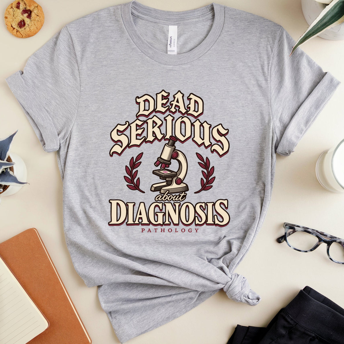 A heather gray graphic t-shirt with a distressed design that reads 'DEAD SERIOUS ABOUT DIAGNOSIS PATHOLOGY' around a microscope illustration, styled in a flat lay with a notebook, glasses, plant, and other desk accessories on a beige background.