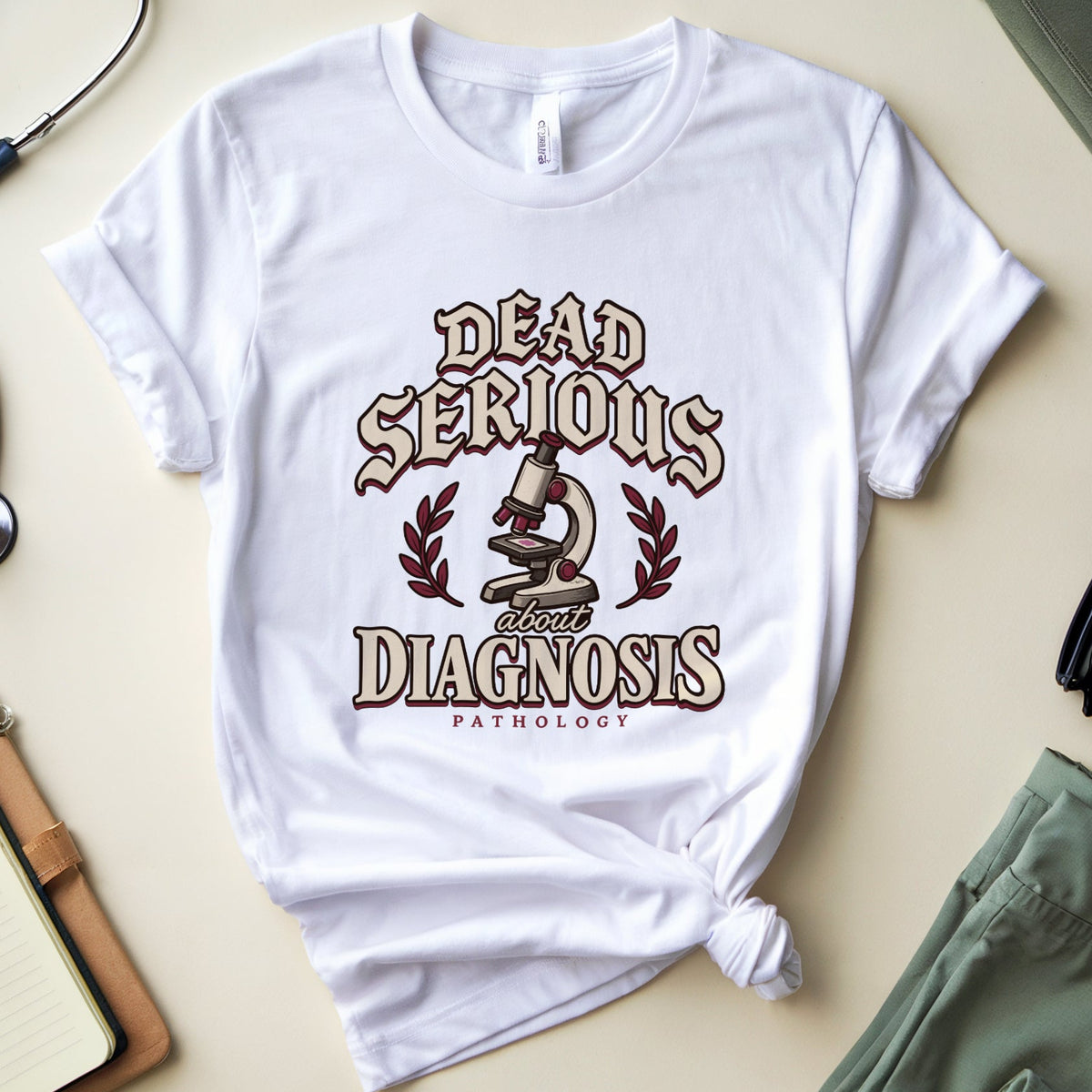 A white graphic t-shirt with a knotted hem, featuring a vintage-style medical design that reads 'Dead Serious Diagnosis' with a microscope and snake motif. The t-shirt is styled in a flat lay with a stethoscope, notebook, glasses, and medical pants on a beige background.