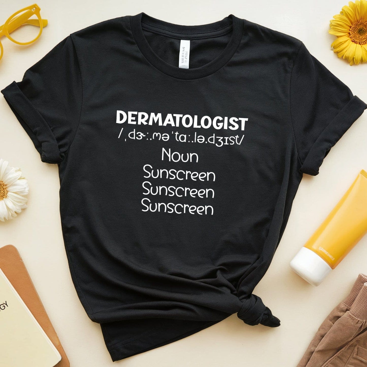 Black t-shirt with 'DERMATOLOGIST' text on a light surface with suncream and flowers.