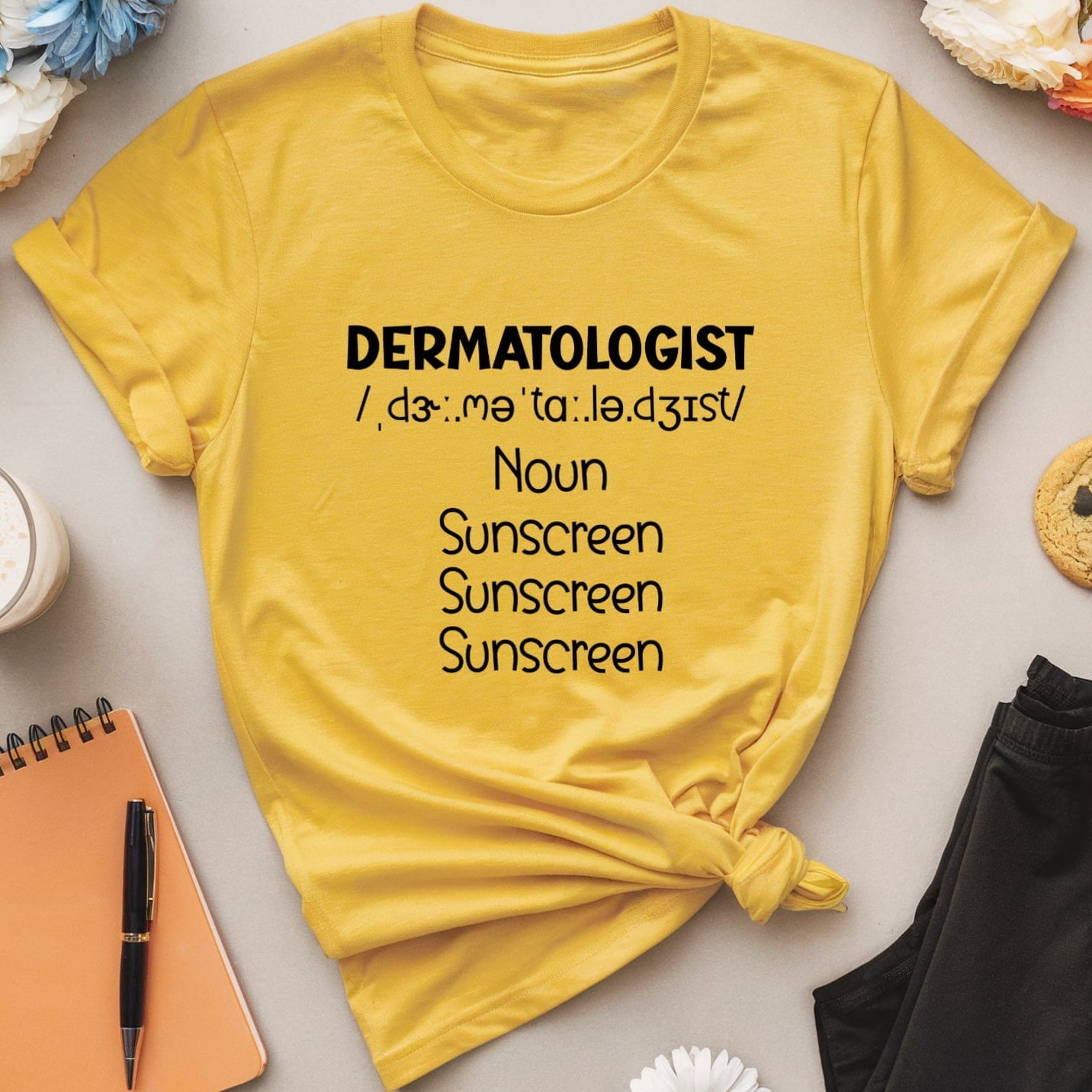Yellow t-shirt with 'DERMATOLOGIST' text on a table with flowers and snacks