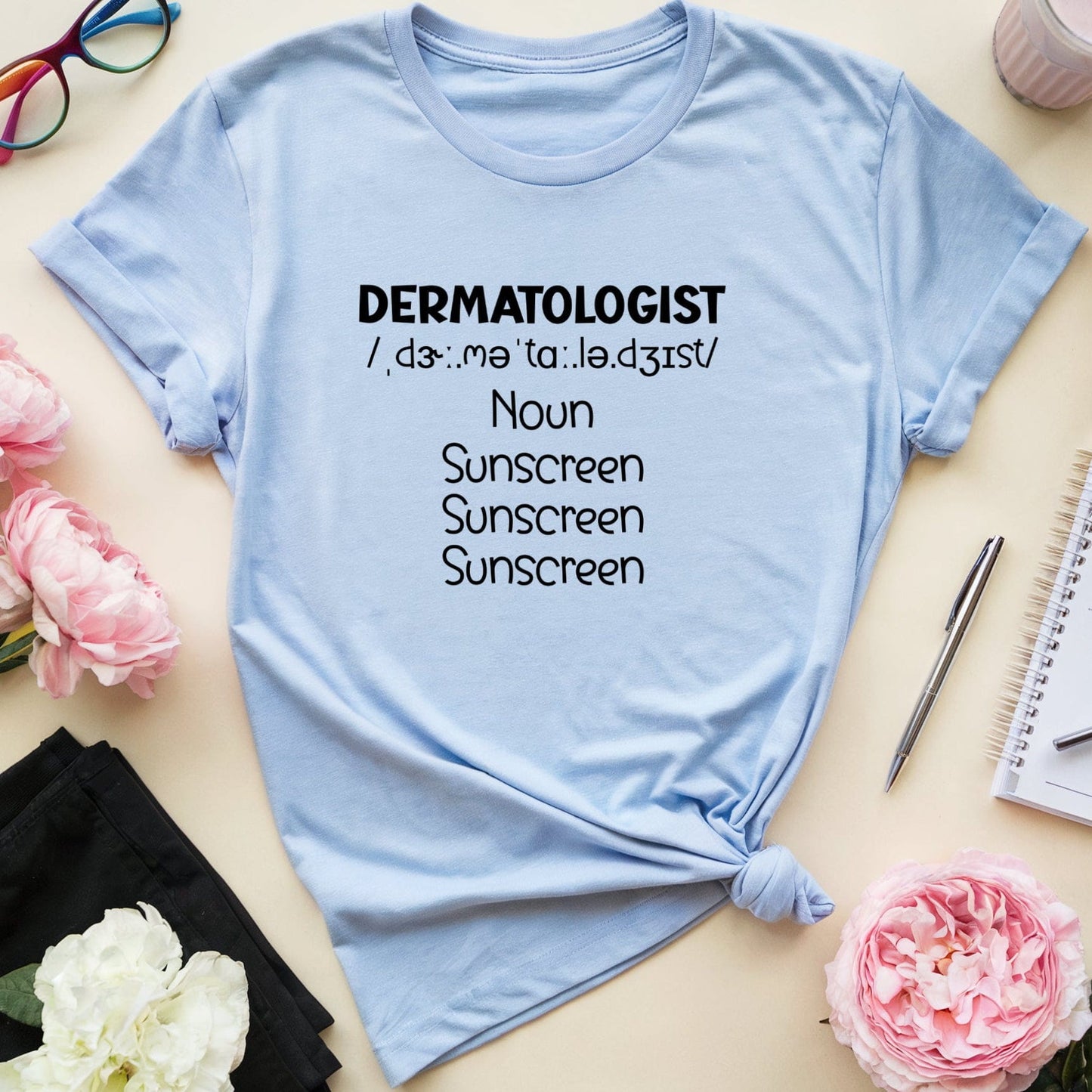 Light blue t-shirt with 'DERMATOLOGIST' definition text on a table with flowers and glasses.