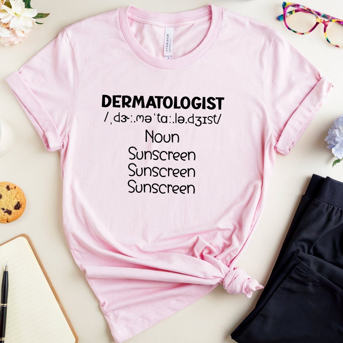 Pink t-shirt with 'DERMATOLOGIST' text on a light surface with accessories.