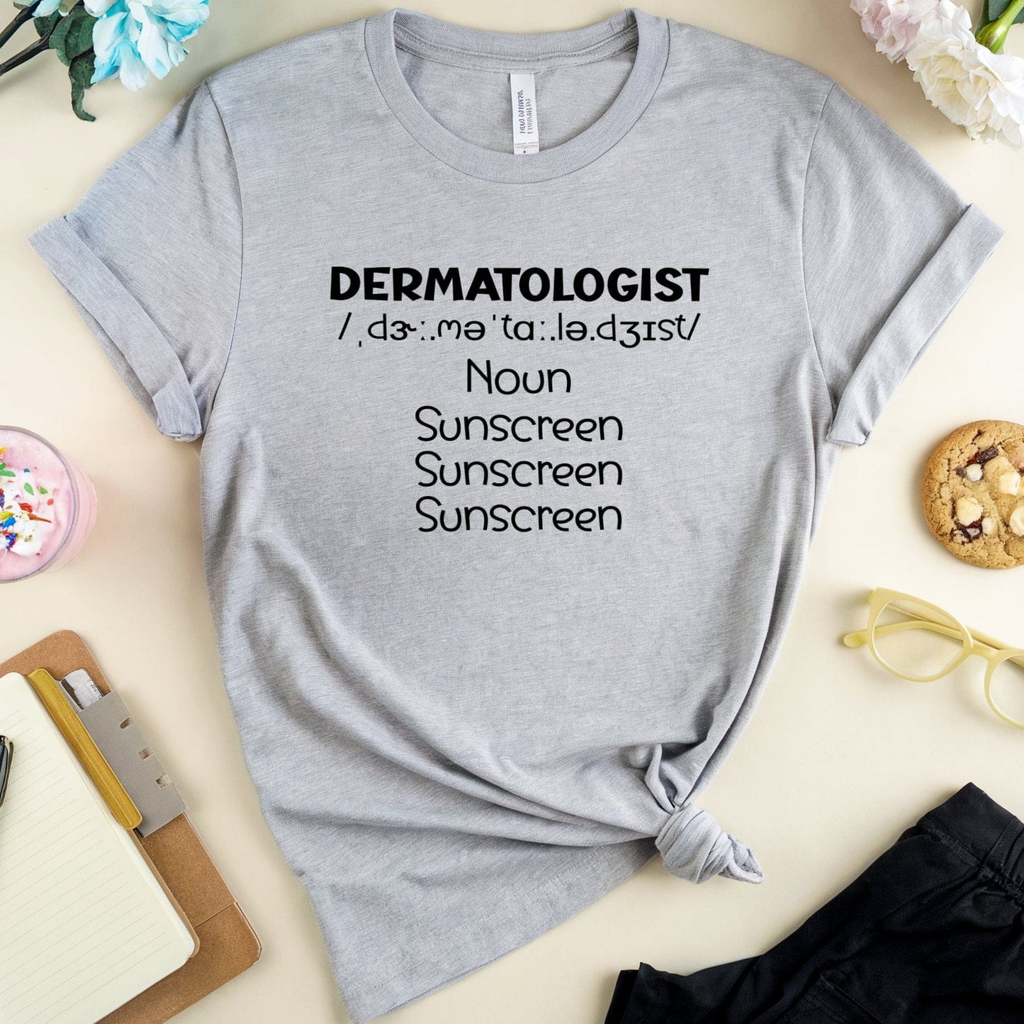 Gray t-shirt with 'DERMATOLOGIST' definition text on a light surface with accessories.