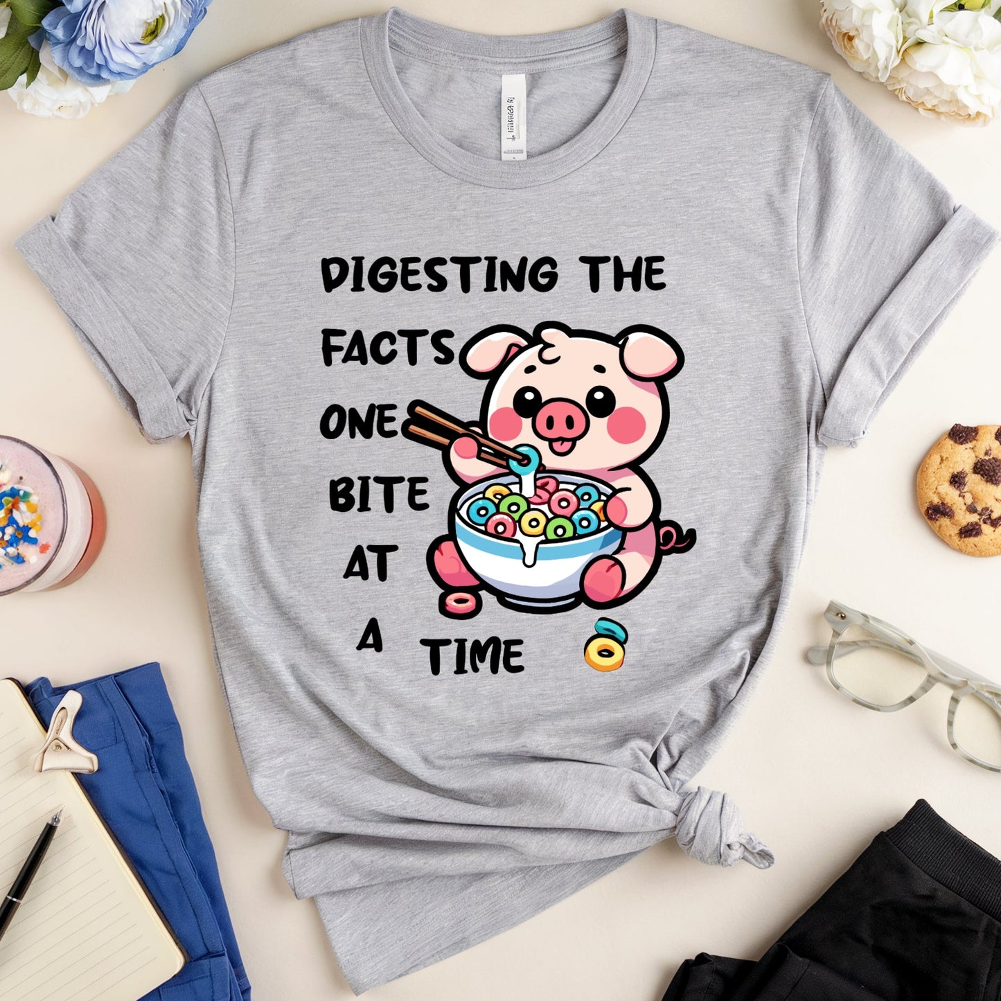 Gray t-shirt with a cartoon pig eating cereal and text, placed on a surface with accessories.