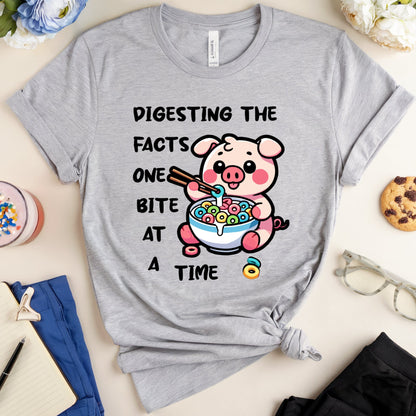 Gray t-shirt with a cartoon pig eating cereal and text, placed on a surface with accessories.